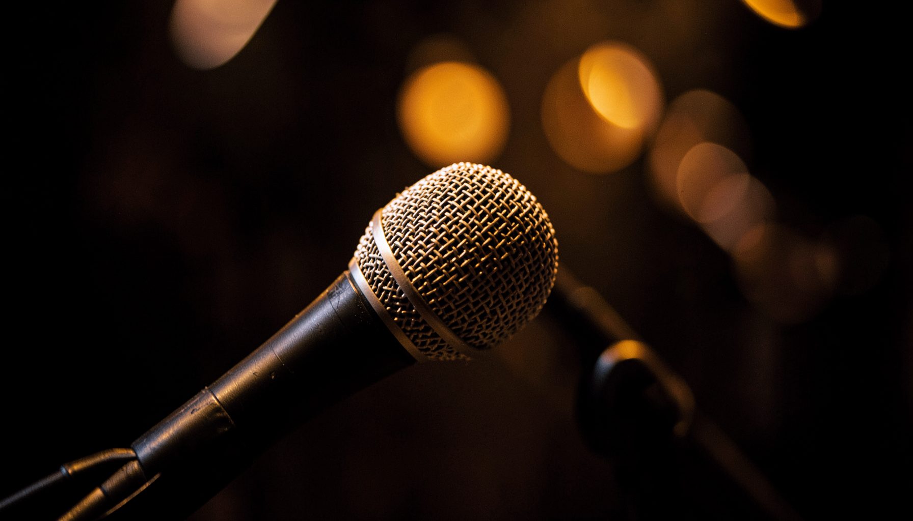 A close-up of a microphone on a dark stage, warm amber and deep brown tones, soft bokeh lights in the background.