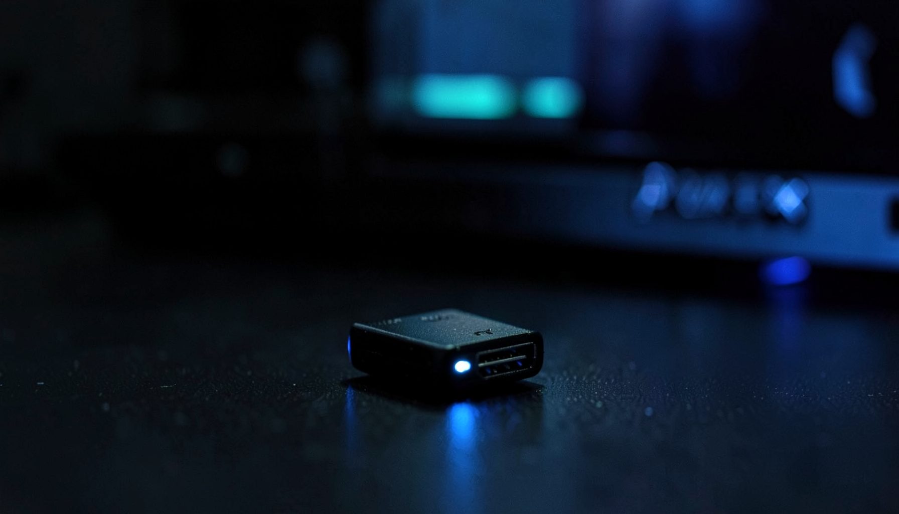 A close-up of a small black USB dongle resting on a dark surface, with subtle blue LED glow and a PlayStation 3 console blurred in the background. Moody, tech-focused atmosphere with deep blacks and cool blue accents.
