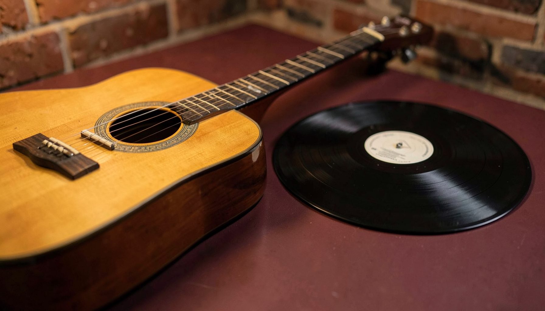 Close view of an acoustic guitar and vinyl record on a muted maroon tabletop, warm brick-toned lighting