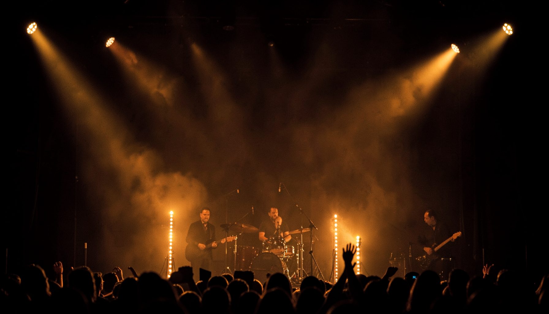 Warm amber stage lights glowing against a dark concert backdrop, with a subtle haze and rich golden-brown tones evoking an intimate live music atmosphere.